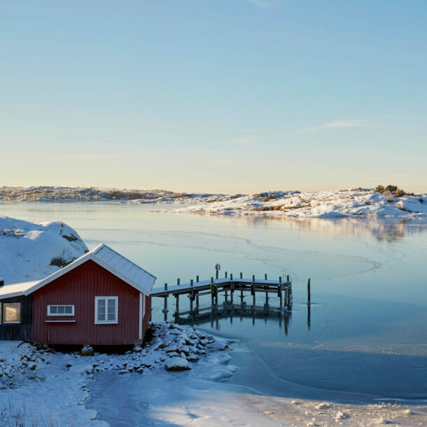 Ein rotes Holzhaus mit Steg an einem zugefrorenen See in verschneiter Landschaft in Schweden.