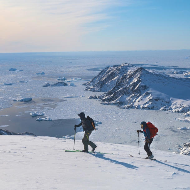 Drei Personen auf Skiern laufen einen schneebedeckten Bergkamm vor einer Gletscherlandschaft entlang.