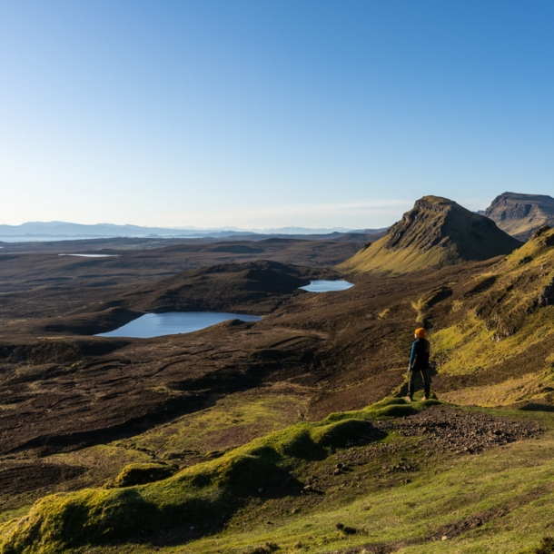 Eine Person steht in einer Landschaft aus Wiesen, Hügeln und Seen in Schottland. 