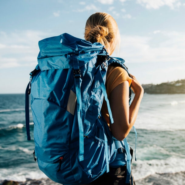 Rückansicht einer Frau mit großem blauen Rucksack am Strand, die aufs Meer blickt.