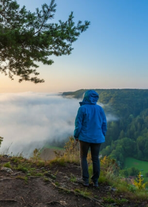 Frau in blauer Funktionsjacke blickt in ein Tal voller Nebel