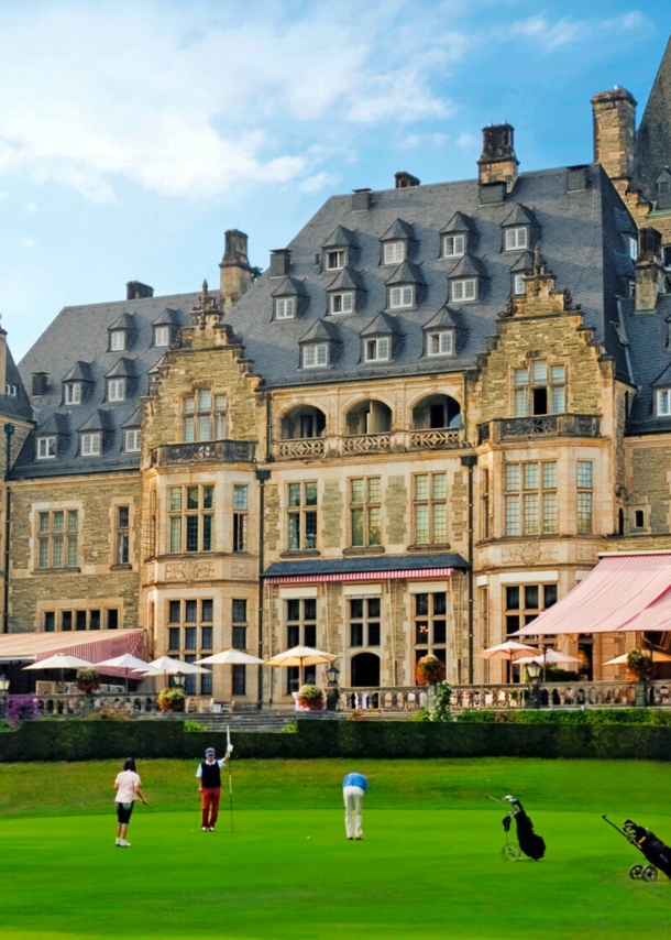 Außenaufnahme von Schlosshotel mit Terrasse, davor Rasen mit Golfenden