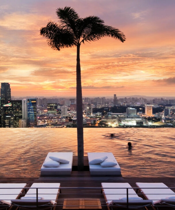 Ein endlos scheinender Pool mit Blick auf die Skyline Singapurs, mit Liegen und Palmen im Wasser