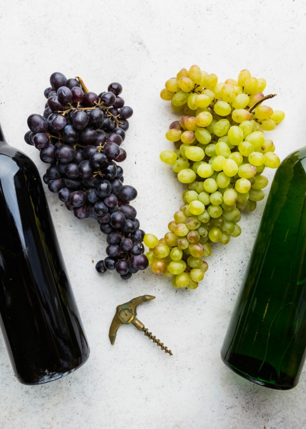 Je eine Flasche und ein Glas Rotwein und Weißwein auf einer hellen Fläche, dazwischen liegen Trauben.