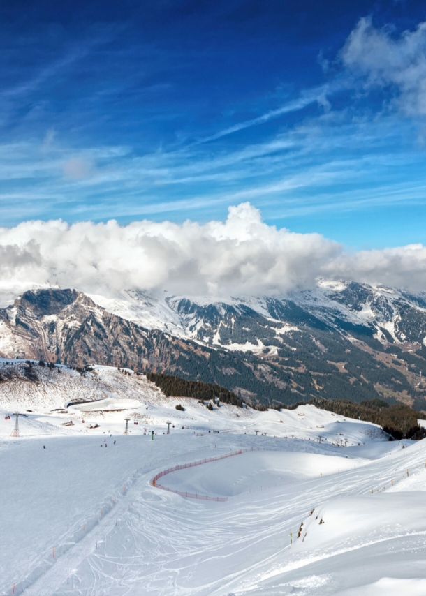 Blick von oben auf eine Piste mit Skifahrer:innen, im Hintergrund Berge