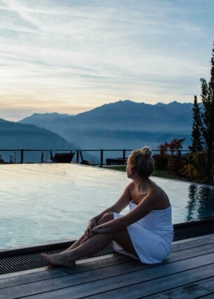 Eine Frau mit umwickelten Handtuch sitzt an einem Outdoor-Pool mit Blick auf die Berge in der Dämmerung