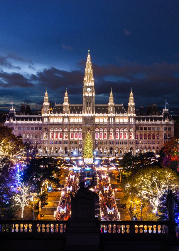 Panoramablick auf das hell erleuchtete Wiener Rathaus und den Rathausvorplatz mit bunt beleuchteten Bäumen bei Nacht