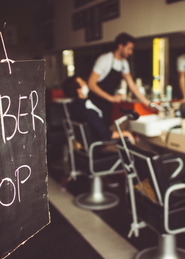 Eine Tafel auf der mit Kreide Barbershop steht, im Hintergrund ist ein Barbier mit seinem Kunden im Laden zu sehen