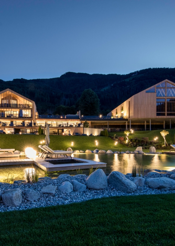 Eine luxuriöse, beleuchtete Hotelanlage aus Holzgebäuden mit Landschaftsgarten und Badeteich vor Bergpanorama bei Nacht