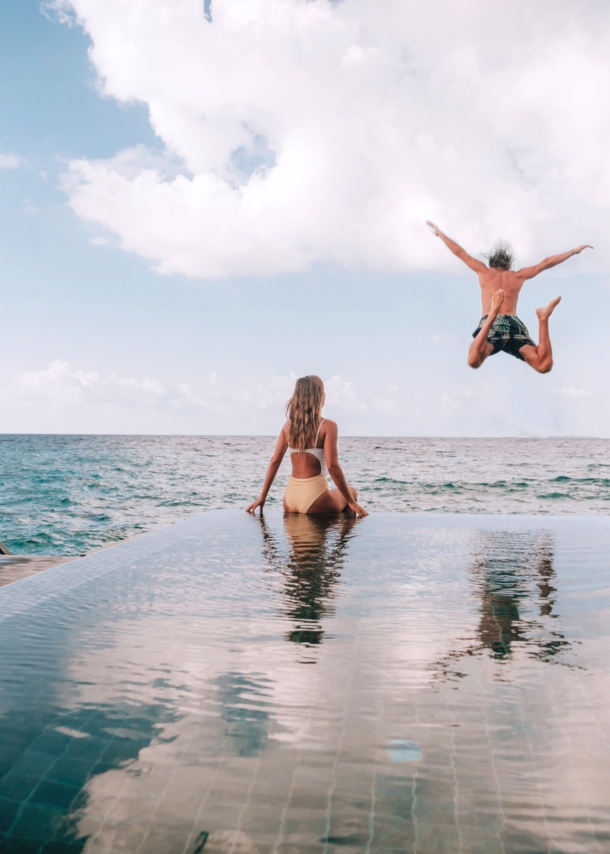 Eine Frau sitzt am Rande eines Pools am Meer, während ein Mann ins Meer springt