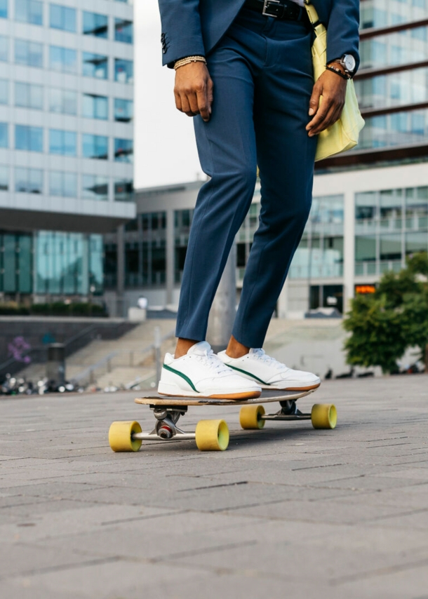 Zwei Beine auf einem Skateboard, die Person trägt eine Anzughose und weiße Sneaker