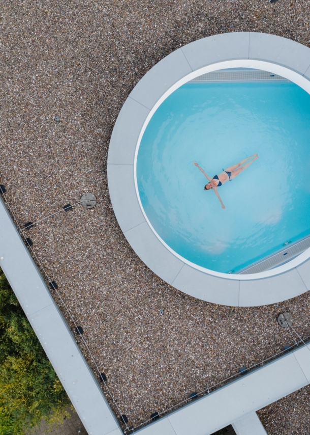 Eine Frau schwebt mit ausgestreckten Armen in einem Pool, fotografiert durch ein rundes Dachfenster aus der Vogelperspektive