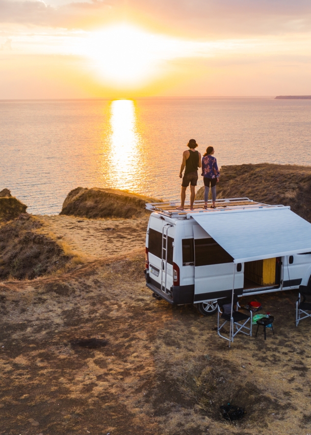 Ein Paar auf dem Dach eines Wohnmobils mit Blick auf die tiefstehende Sonne über dem Meer