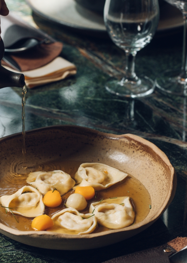 An einem stilvoll gedeckten Restauranttisch läuft Brühe aus einer Kanne in einen Teller mit gefüllter Pasta