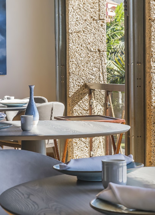 Innenraum eines luftigen, hellen Restaurants mit gedeckten Tischen an einer Fensterfront mit Blick auf Palmen