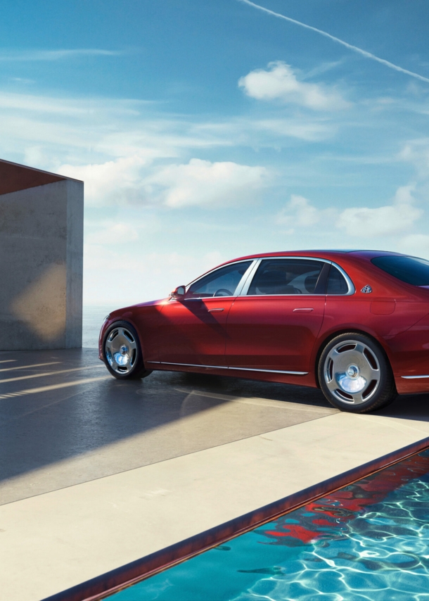 Ein roter Mercedes-Maybach von der Seite am Pool eines modernen Hauses, im Hintergrund Meer und Personen im Haus