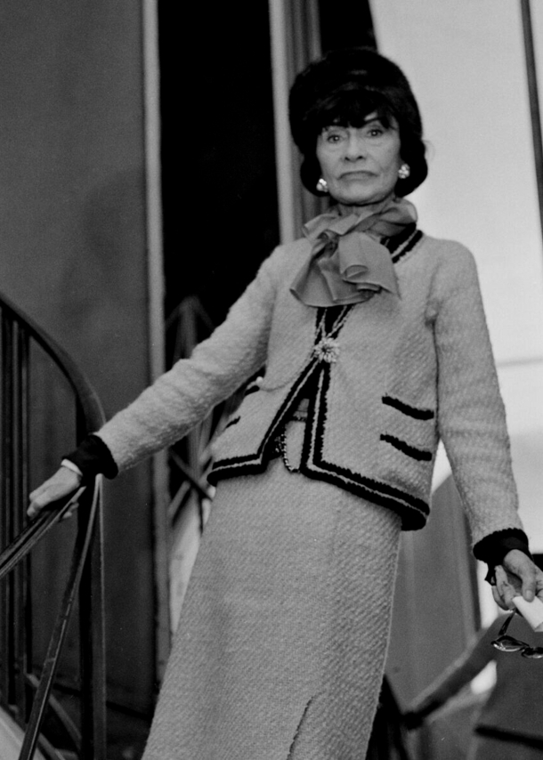 Schwarzweißaufnahme von Coco Chanel in einem Tweed-Kostüm auf einer Treppe vor einer Spiegelwand