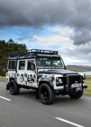 Der Defender Works V8 Trophy II fährt bei bewölktem Wetter an einem See entlang.