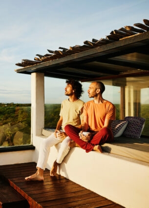 Zwei lässig gekleidete Männer sitzen auf einer Terrasse in der Abendsonne und schauen über eine tropische Landschaft aufs Meer