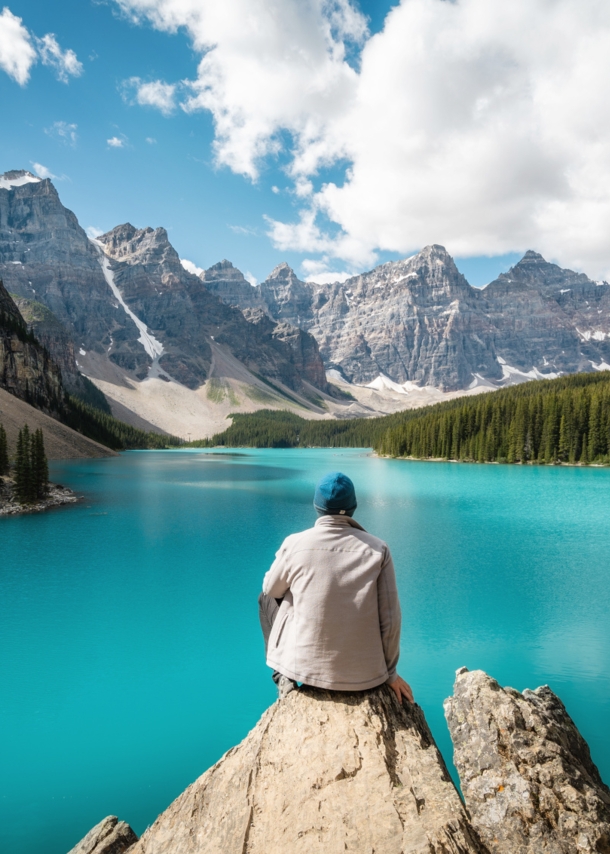 Eine Person sitzt auf einem Stein und schaut auf einen See, der gesäumt von bewaldeten Bergen ist