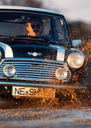 Ein Mann fährt in einem schwarzen MINI Cooper 1.3 mit Rallystreifen durch eine Schlammpfütze