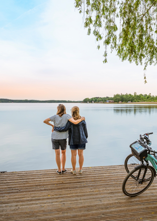 Rückansicht von zwei Frauen, die auf einem Steg vor einem See stehen und sich umarmen, daneben zwei E-Bikes