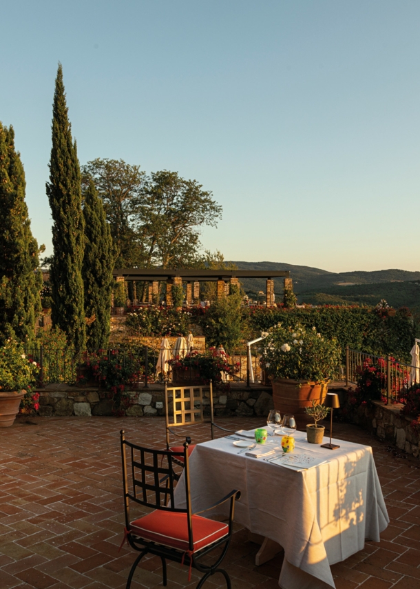 Ein gedeckter Tisch auf einer großen Terrasse einer Villa im toskanischen Baustil mit Blick in eine grüne Hügellandschaft bei Sonnenuntergang