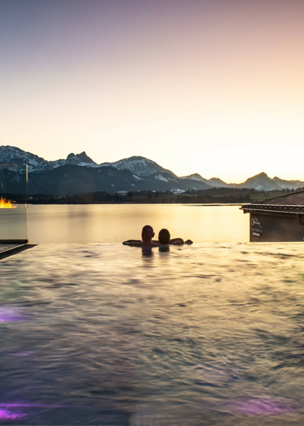 Zwei Personen in einem Infinity-Pool schauen über einen See in eine Berglandschaft bei Sonnenuntergang