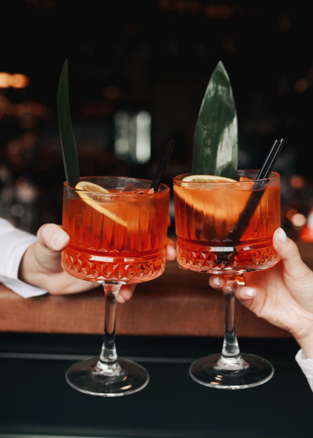 Zwei Spritz-Getränke mit Orangenscheiben in den Händen zweier Personen beim Anstoßen an einem Bartresen.