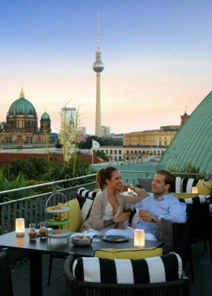 Ein junges Paar sitzt gut gelaunt an einem Tisch auf einer Dachterrasse, im Hintergrund Berliner Dom und Fernsehturm.