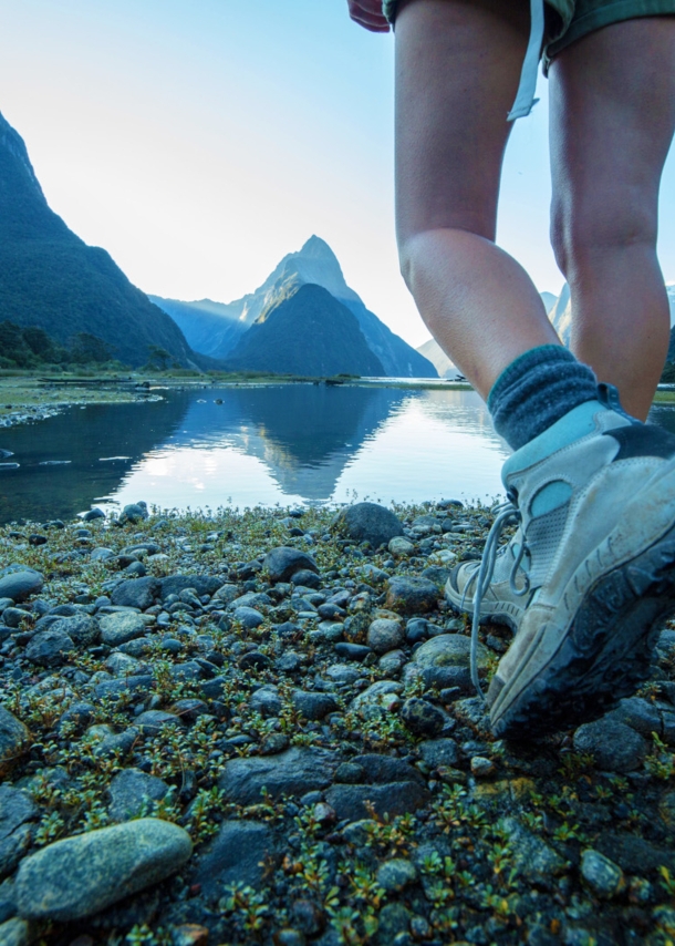 Zwei Beine einer Person in Wanderschuhen an einem See vor Bergpanorama.