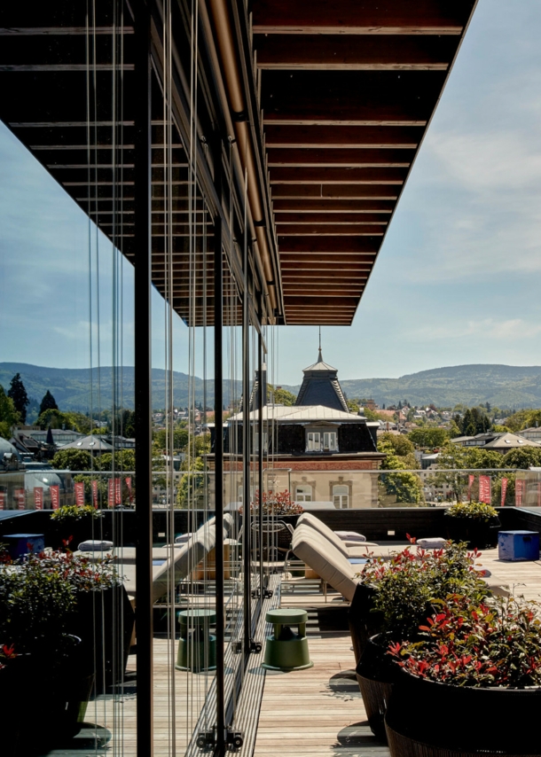 Große Balkonterrasse mit Liegestühlen vor grüner Hügellandschaft, die sich in der Glasfassade des Gebäudes spiegelt.