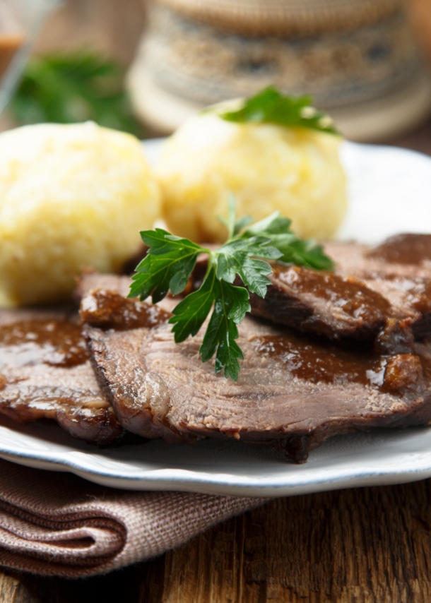 Sauerbraten mit brauner Soße an Klößen, angerichtet auf einem weißen Teller auf einem Holztisch.