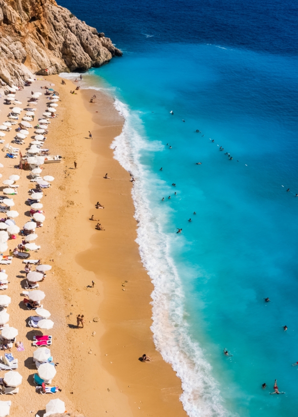 Luftaufnahme eines goldenen Sandstrandes mit Sonnenschirmen in einer Felsbucht mit Personen im türkisblauen Wasser.