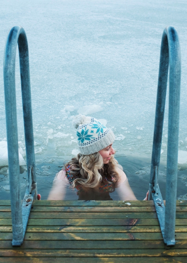 Eine Frau mit Wollmütze hält sich beim Baden in einem zugefrorenen See an dem Handlauf einer Treppe an einem Holzsteg fest.