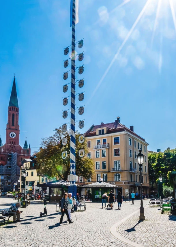 Ein Stadtteilplatz mit Kopfsteinpflaster und einem Maibaum in der Mitte bei strahlendem Sonnenschein.