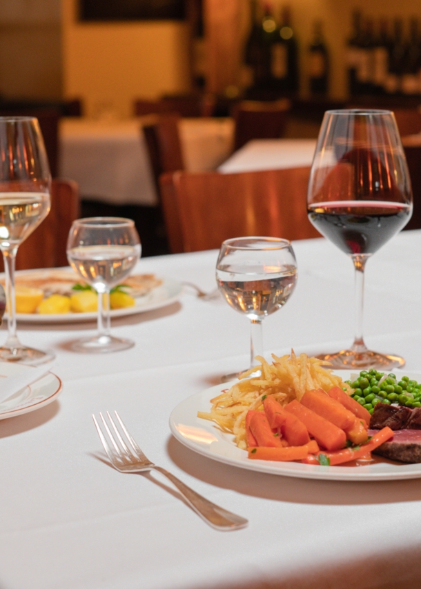 Zwei auf weißen Tellern angerichtete Speisen stehen neben Weingläsern auf einem Tisch mit weißer Stofftischdecke in einer Brasserie.