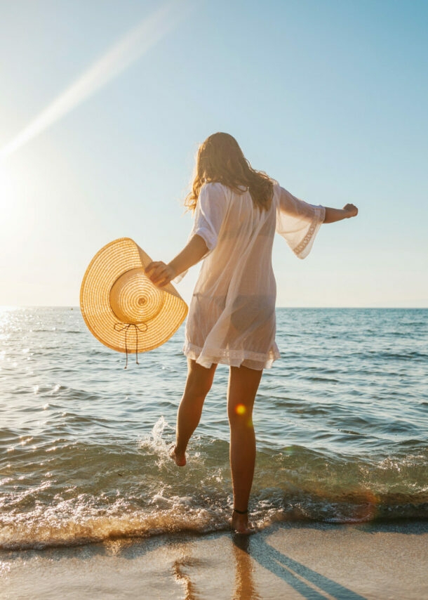 Eine junge Frau im weißen Sommerkleid und mit Strohhut in der Hand planscht mit ihrem Fuß im Wasser an einem Sandstrand am Meer bei Sonnenuntergang