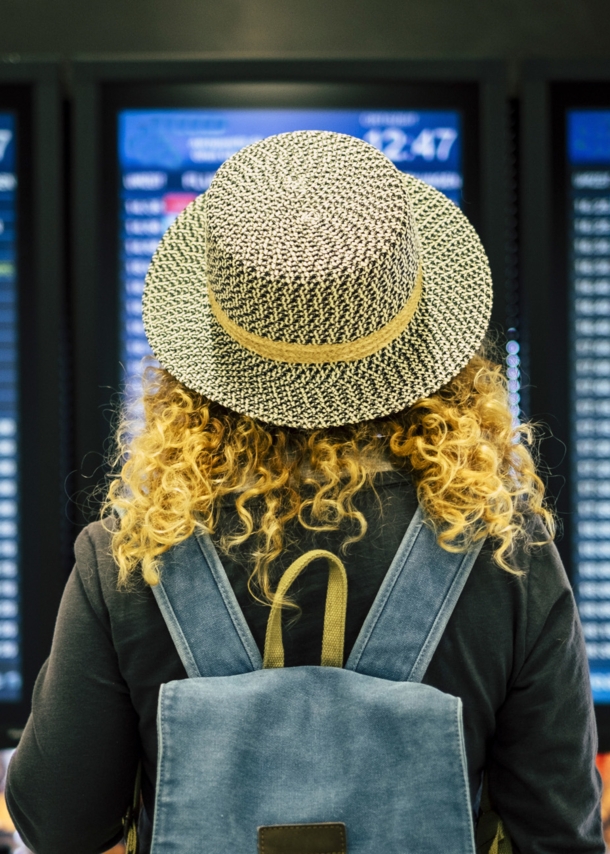 Rückansicht einer Frau mit Strohhut vor einer Anzeigetafel am Flughafen.