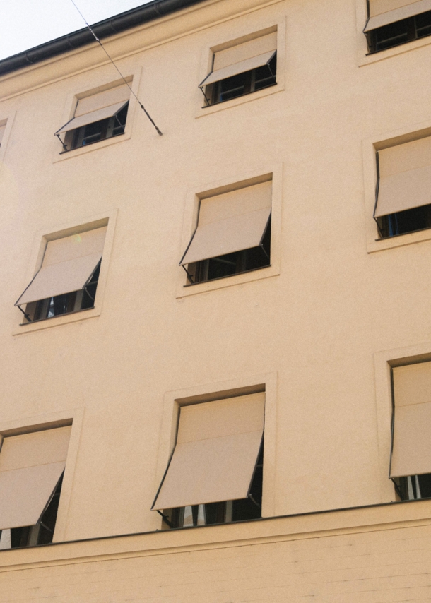 Hellbeige Hausfassade mit gleichfarbigen Markisen vor den Fenstern.