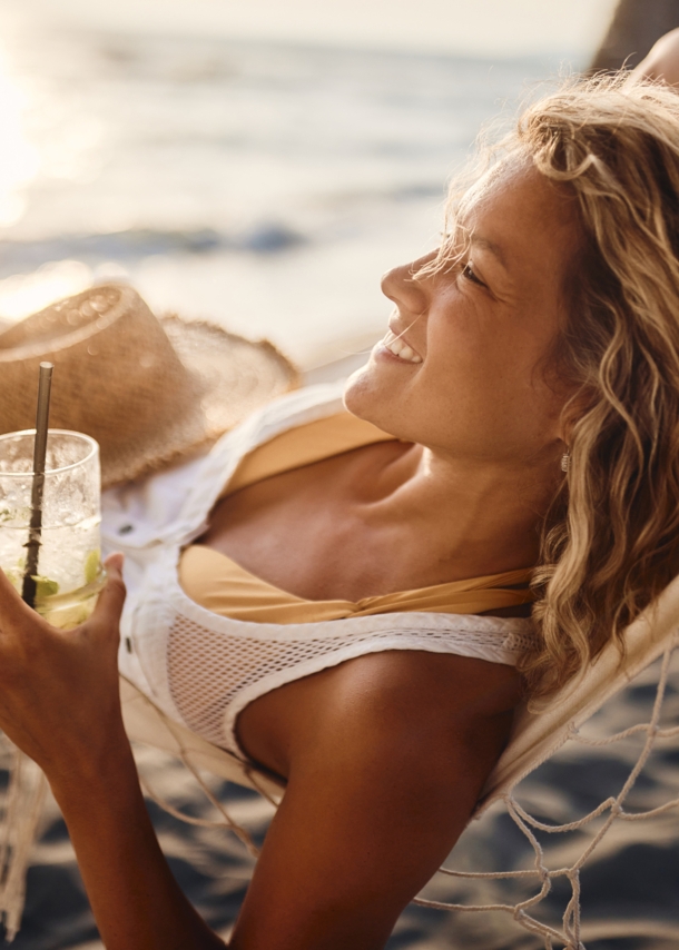 Eine junge Frau liegt entspannt mit einem Getränk in der Hand in einer Hängematte am Strand