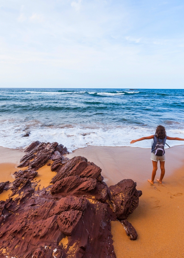 Eine Person steht mit dem Rücken zur Kamera und neben einem Felsen an einem Strand und schaut mit ausgebreiteten Armen aufs Meer