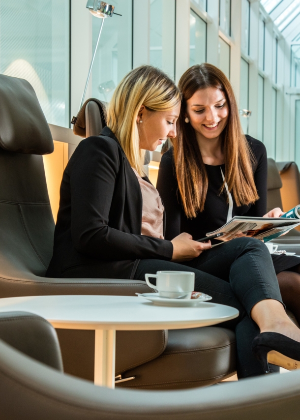 Zwei Frauen auf Ledersesseln schauen gemeinsam auf ein Magazin in einer Lounge.