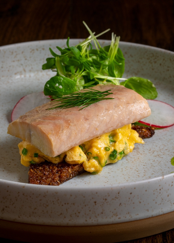 Ein Keramikteller mit hohem Rand, auf dem ein Stück Fisch mit weiteren Zutaten hübsch angerichtet liegen.