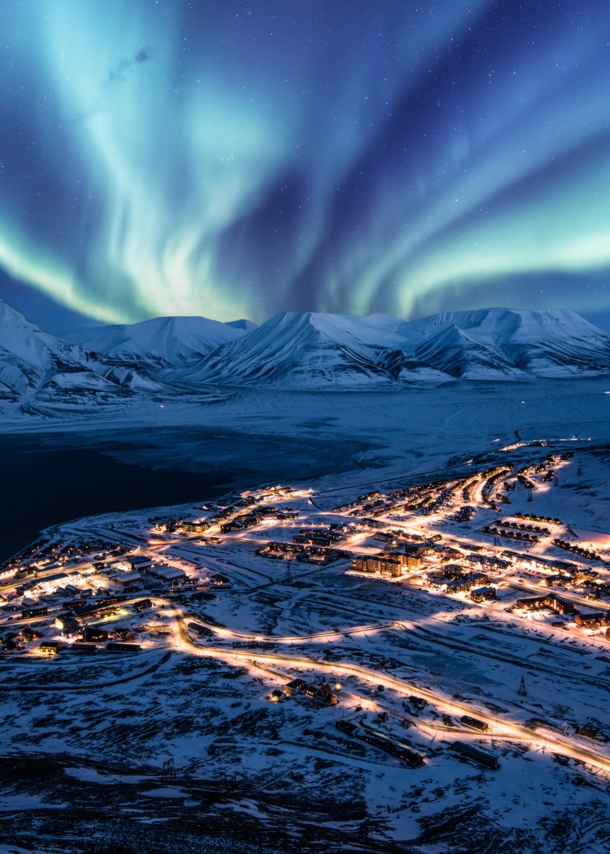 Beleuchtete Ortschaft in schneebedeckter Fjordlandschaft unter Polarlichtern bei Nacht.