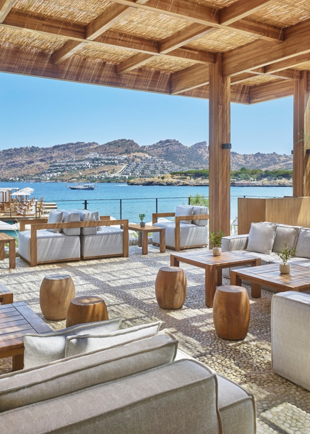 Loungebereich mit grauen Sesseln und Holztischen auf einer überdachten Terrasse an einer mediterranen Bucht.