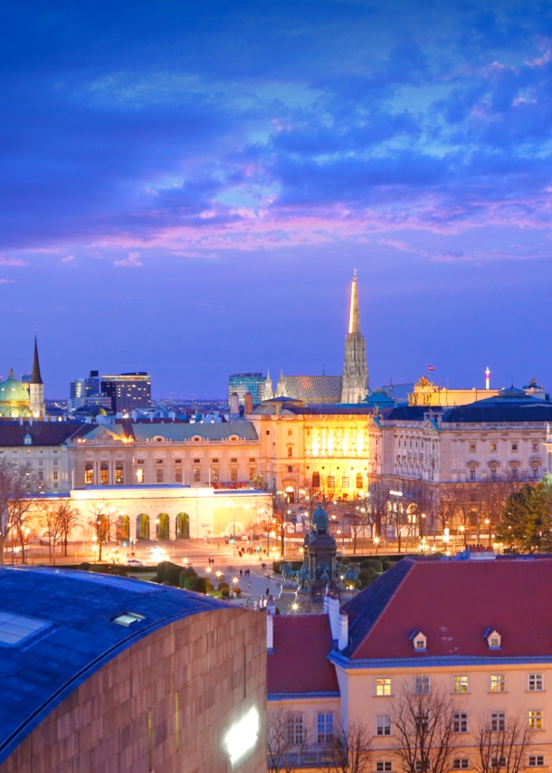 Blick von oben auf eine Stadt in der Dämmerung. Im Vordergrund sind prächtige, beleuchtete Gebäude zu sehen.