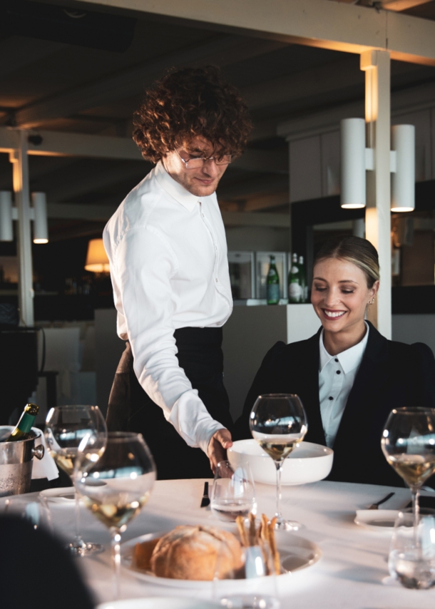 Eine lächelnde Frau wird in einem Restaurant von einem Kellner bedient, der einen Teller vor ihr auf den weiß eingedeckten Tisch stellt.