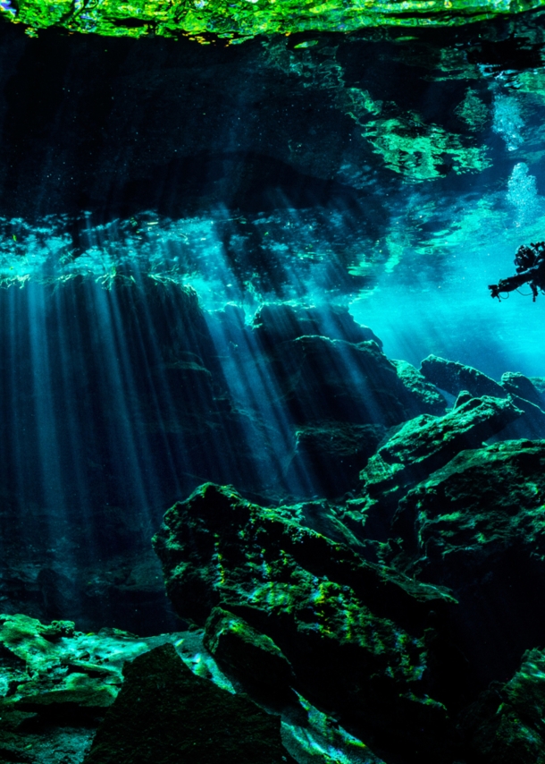 Eine Person in Taucherausrüstung taucht in einer von Wasser gefüllten Höhle. Lichtsäulen ergeben sich durch den starken Lichteinfall von oben im Wasser.