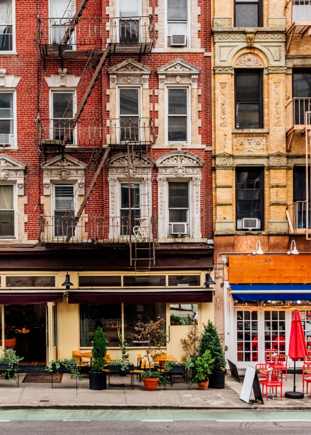 Geschlossene New Yorker Häuserfront mit Feuerleitern und Restaurants mit Außengastronomie.